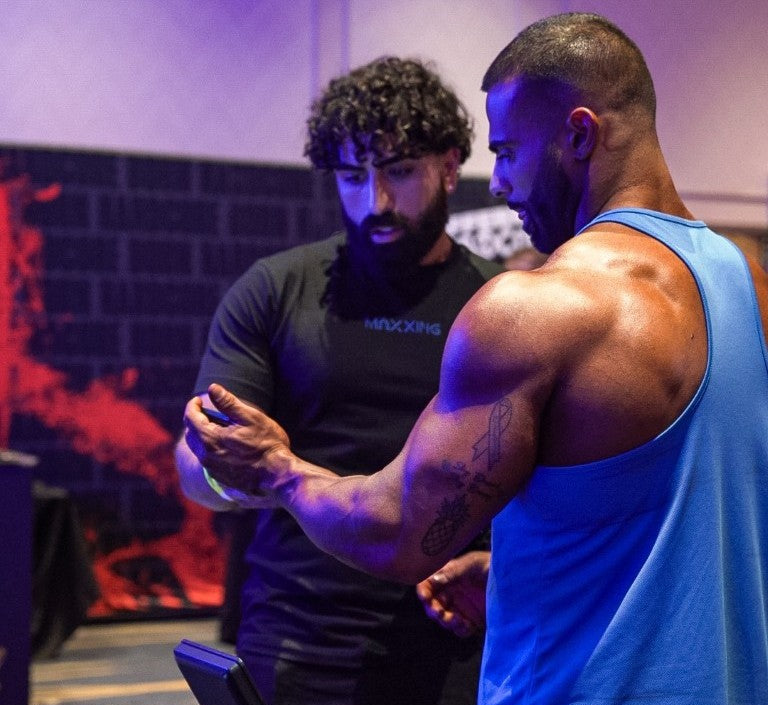 Two men in a room with a dark background, one wearing a black shirt and the other in a blue tank top.