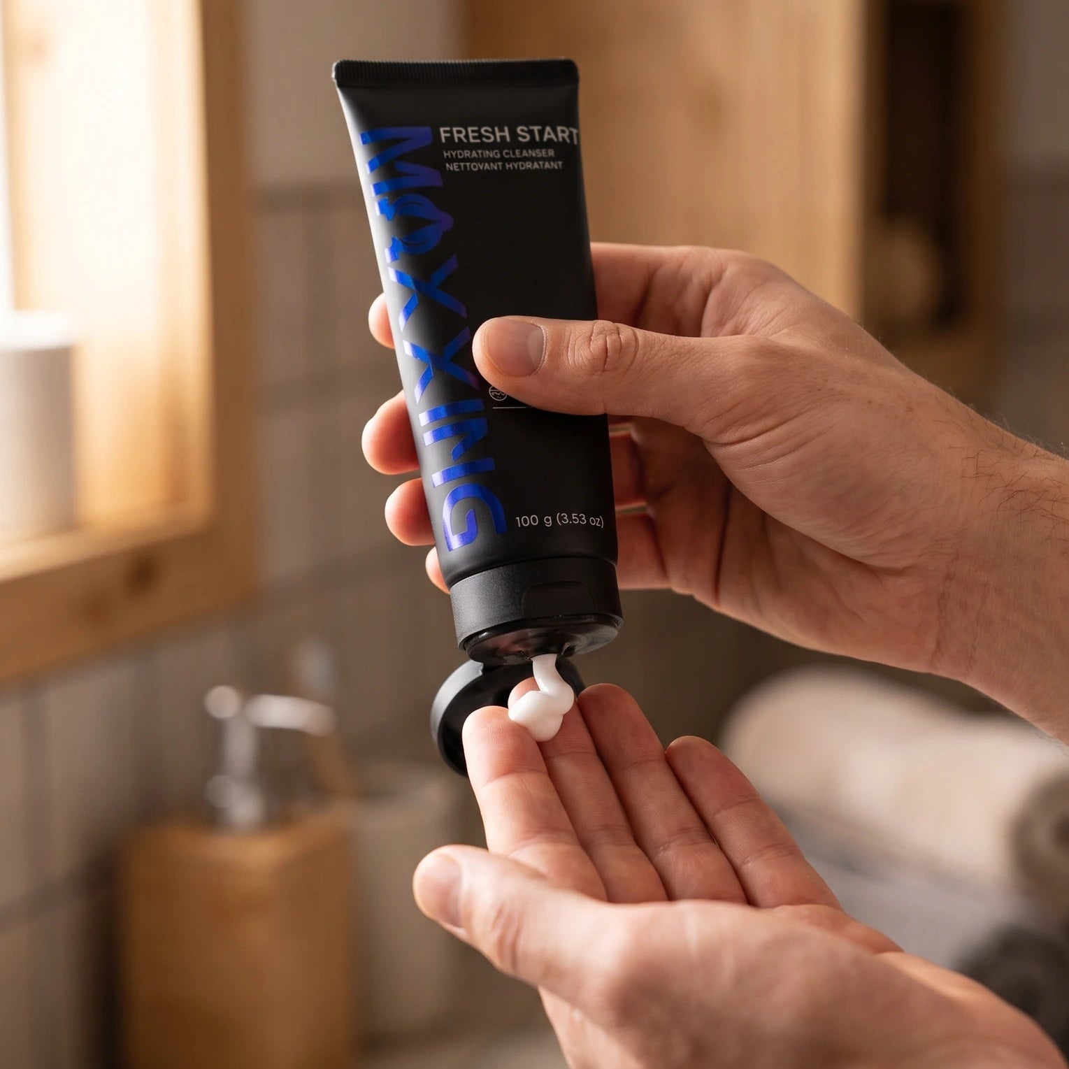 A person's hands squeeze a black tube of hair product labeled 'FRESH START' with blue lettering, dispensing white foam into the other hand in a softly lit bathroom setting.